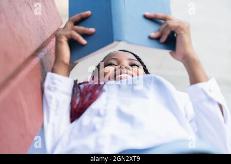 Lächelndes Gesicht einer afrikanischen Frau, die ein Buch liest, während sie sich auf einer Bank im Freien lehnt Stockfoto