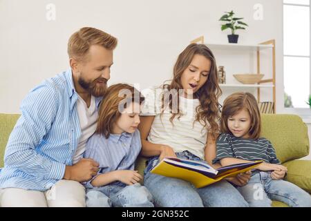 Eltern und kleine Kinder lesen ein Buch mit Geschichten, während sie zu Hause auf der Couch sitzen Stockfoto