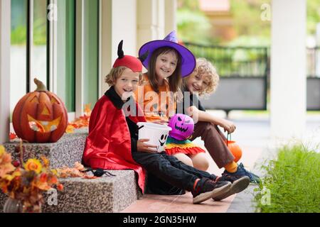 Kinder tricksen oder gönnen sich an Halloween Abend. Kind an der dekorierten Haustür. Junge und Mädchen in Hexe- und Vampir-Kostüm und Hut mit Süßigkeiten Eimer und Kürbis Stockfoto