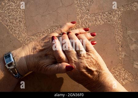 Crop unkenntlich ältere Frau streckt die Hände mit roten Maniküre und goldenem Ring gegen beige Wand im Sonnenlicht Stockfoto