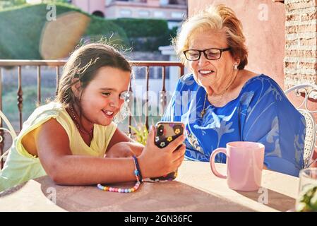 Glückliche Enkelin sitzt am Tisch und zeigt lächelnde Großmutter Fotos auf dem Smartphone, während sie an sonnigen Tagen auf der Terrasse sitzt Stockfoto