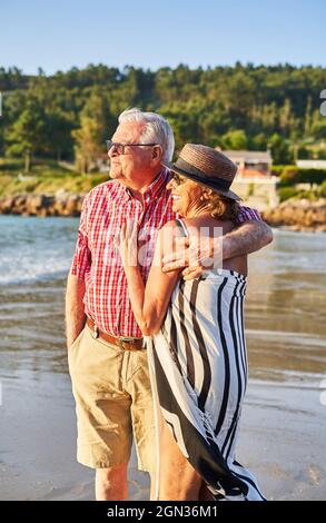 Lächelndes, barfuss gelauntes, älteres Paar in Sonnenbrillen, das sich am nassen Sandstrand umarmt und einen sonnigen Tag genießt Stockfoto