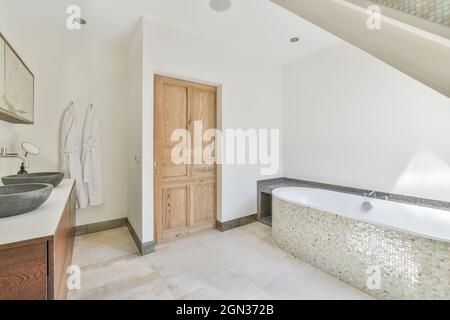 Badewanne mit Mosaikfliesen und Waschbecken zwischen Schränken und Spiegeln im modernen Badezimmer an sonnigen Tagen Stockfoto