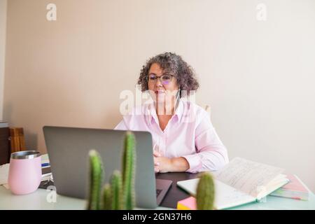 Fokussierte Geschäftsfrau in Brillen und Ohrhörern, die mit Netbook am Tisch sitzt Stockfoto