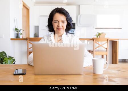 Glückliche, reife Freiberuflerin, die im Internet auf Netbook surfen und an einem neuen Projekt arbeiten kann, während sie zu Hause am Tisch sitzt Stockfoto