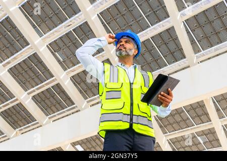 Niedriger Winkel eines männlichen hispanischen Ingenieurs mittleren Alters in Uniform mit Tablet, das unter dem Solarkraftwerk wegschaut Stockfoto