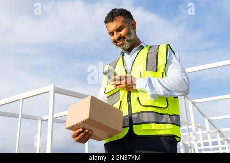 Von unten angenehmer bärtiger ethnischer Arbeiter in Uniform, der das Paket trug, während er bei der Arbeit das Mobiltelefon benutzte Stockfoto