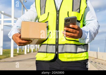 Unbekennbarer männlicher Arbeiter in Uniform, der das Paket trug, während er das Mobiltelefon bei der Arbeit benutzte Stockfoto