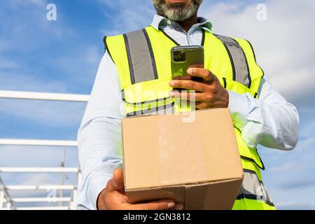 Unbekennbarer männlicher Arbeiter in Uniform, der das Paket trug, während er das Mobiltelefon bei der Arbeit benutzte Stockfoto