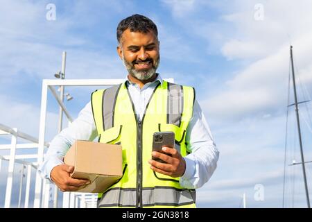 Von unten angenehmer bärtiger ethnischer Arbeiter in Uniform, der das Paket trug, während er bei der Arbeit das Mobiltelefon benutzte Stockfoto