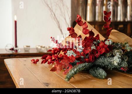 Festlicher stilvoller dekorativer Weihnachtsstrauß mit Zweigen aus Eukalyptus und leuchtend roten Zweigen mit Beeren auf Holztisch im Zimmer Stockfoto