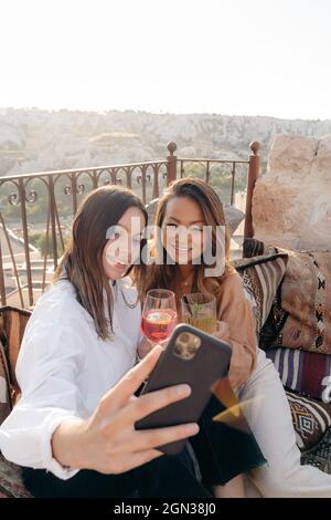 Ein großer Winkel von ethnischen, glücklichen Freundinnen, die auf Kissen sitzen und sich mit einer Sofortbildkamera und einem Cocktail auf dem Handy neben dem Tisch porträtieren Stockfoto