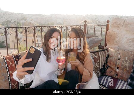 Ein großer Winkel von ethnischen, glücklichen Freundinnen, die auf Kissen sitzen und sich mit einer Sofortbildkamera und einem Cocktail auf dem Handy neben dem Tisch porträtieren Stockfoto