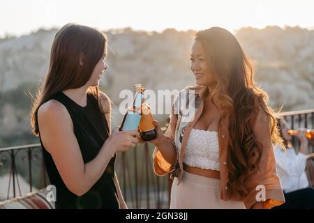 Seitenansicht positiver junger multiethnischer Freundinnen, die hell lächeln und klirrende Bierflaschen trinken, während sie beim Sonnenuntergang eine angenehme Zeit zusammen genießen Stockfoto