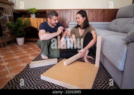 Mann, der eine Schraube an eine fröhliche asiatische Frau hergab, während er im Wohnzimmer einen Tisch auf einem Zierteppich montierete Stockfoto