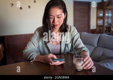Junge, ethnische Frau mit einem Glas Kaffee, die am Tisch im Zimmer im Internet surfen konnte Stockfoto