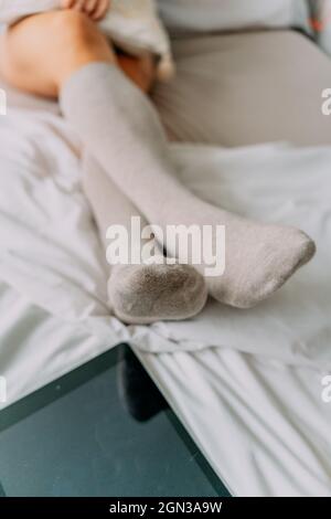 Crop unkenntlich weiblich in Kniesocken sitzen mit gekreuzten Beinen gegen Handy mit schwarzem Bildschirm auf zerknittertem Bettlaken Stockfoto