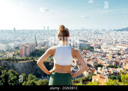 Rückansicht einer anonymen Athletin mit Händen an der Taille, die die Sommerstadt im Sonnenlicht bewundert Stockfoto