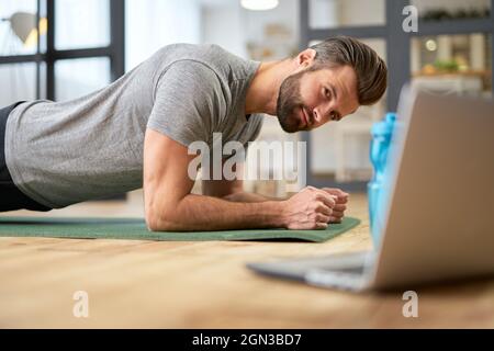 Bärtiger junger Mann, der zu Hause ein Notebook benutzt und eine Planke macht Stockfoto