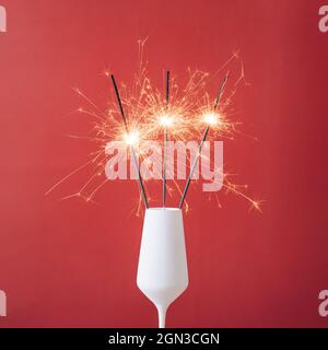 Nahaufnahme von drei Wunderkerzen in einem weißen Glas Champagner vor rotem Hintergrund für einen Kopierraum als Symbol der Freude an der Feier des neuen Jahres Stockfoto