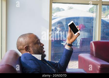 Mann in einem blauen Business-Anzug sitzt auf dem Sessel und hält sein Telefon. Stockfoto