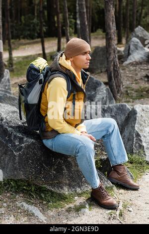 Reisende mit Rucksack, in warmer Weste und Mütze, sitzend auf Stein im Wald Stockfoto