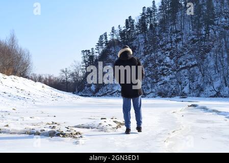 Mann in warmen Kleidern auf gefrorenem Fluss im Wald stehen und Blick auf verschneite Berge. Winter, Russland. Stockfoto