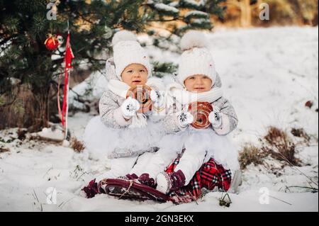 Zwei kleine Zwillingsmädchen in weißen Anzügen sitzen und essen im Winter draußen große Brötchen. Stockfoto