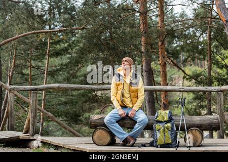 Reisende blicken weg, während sie sich auf einer Holzbank in der Nähe von Rucksack und Trekkingstöcken ausruhen Stockfoto