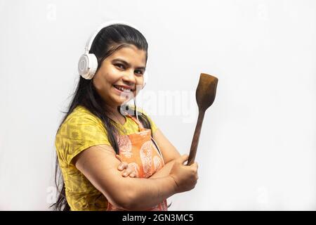 Eine hübsche indische Hausfrau, die Kochschürze und Kopfhörer mit Holzspatel in der Hand trägt, lächelt auf weißem Hintergrund Stockfoto