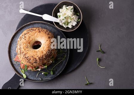 Teller mit Bagel mit Frischkäse, trockenem Schinken und Microgreens auf grauem Hintergrund, köstliches Snackkonzept, Draufsicht. Stockfoto