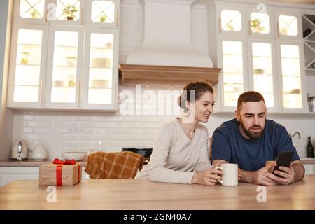 Fröhliches junges kaukasisches Paar, das in der Küche am Tisch sitzt und Kaffee trinkt, während es auf dem Smartphone gemeinsam im Internet surft Stockfoto