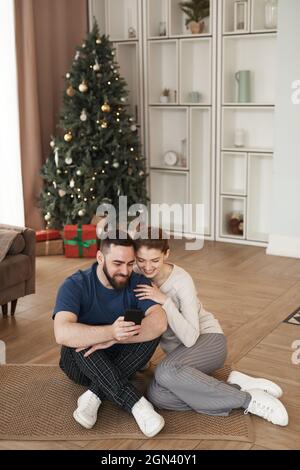 Lächelnder junger bärtiger Mann, der im Wohnzimmer mit Weihnachtsbaum auf dem Boden sitzt und der Freundin ein Video am Telefon zeigt Stockfoto