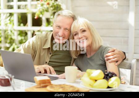 Glückliches Seniorenpaar, das im Freien einen Laptop benutzt, auf der Terrasse sitzt und vor der Kamera lächelt Stockfoto