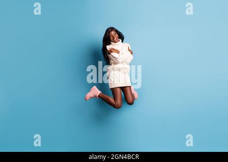 In voller Länge Foto von niedlichen dunklen Haut Frau gekleidet weißen Winterkleidung springen umarmt sich isoliert blauen Hintergrund Stockfoto