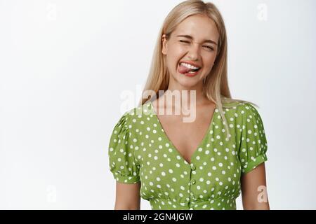 Lustige blonde Frau zeigt Zunge und lächelt, zwinkert fröhlich an der Kamera, steht vor weißem Hintergrund in grünem Kleid Stockfoto