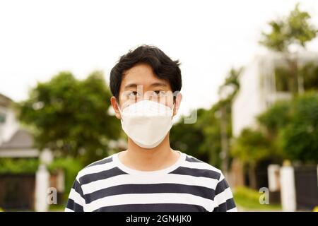 Porträt eines jungen, hübschen asiatischen Mannes mit covid-19 Gesichtsschutz-Sicherheitsmaske, der tagsüber im Freien steht und die Kamera anschaut Stockfoto