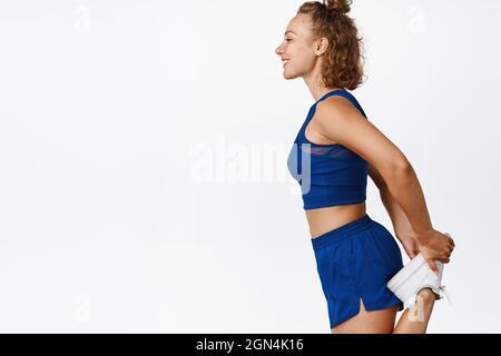 Profilaufnahme einer Fitness-Frau, die beim Joggen des Warm-Up ihr Bein streckt. Fröhliche Sportlerin, die ihr Workout macht, läuft in Active-Ear, weiß Stockfoto