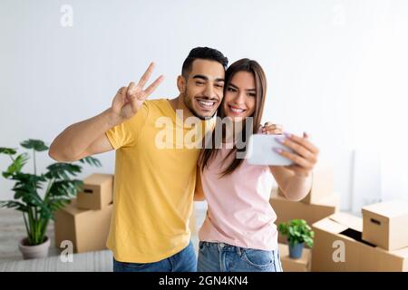 Lächelnder Araber und seine kaukasische Freundin ziehen zusammen in ihr eigenes Zuhause und machen Selfie zwischen Kartons Stockfoto