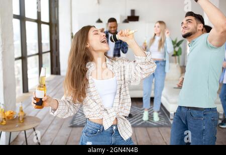 Junge blonde Dame feiert mit ihren vielfältigen Freunden, singt Karaoke ins Mikrofon, tanzt und trinkt zu Hause Bier Stockfoto