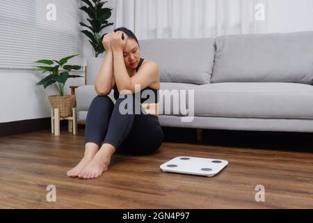 Junge asiatische Frau, die auf dem Boden sitzt und die Waage anschaut, ist besorgt und deprimiert, wenn es um Diät- und Essstörungen geht Stockfoto