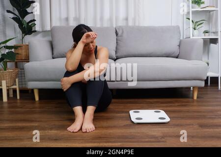 Junge asiatische Frau, die auf dem Boden sitzt und die Waage anschaut, ist besorgt und deprimiert, wenn es um Diät- und Essstörungen geht Stockfoto