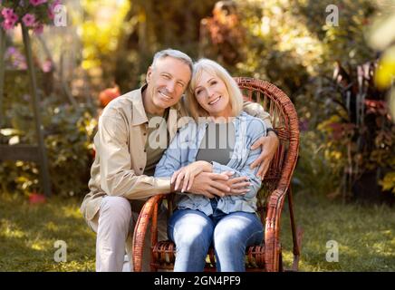 Liebende ältere Ehepartner ruhen auf dem Land, Frau sitzt in Korbstuhl, Mann umarmt Frau, kopieren Raum Stockfoto