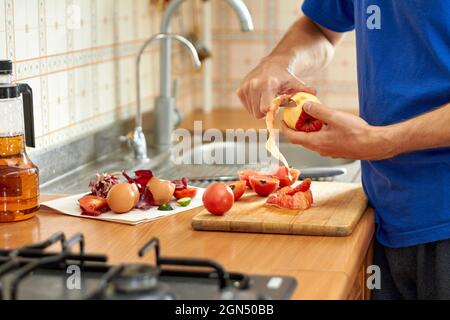 Ein Mann schneidet eine Schale mit einem Messer aus einem roten Apfel. Lebensmittelabfälle und Reste während der Zubereitung von Speisen in der Küche. Nahaufnahme, selektiver Fokus Stockfoto