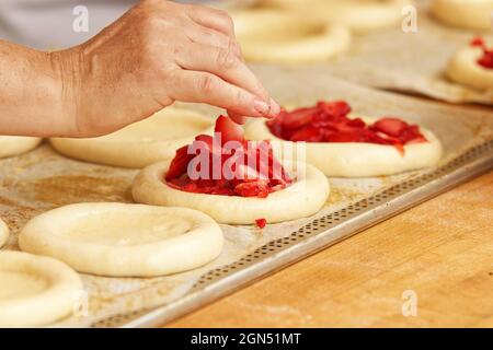 Die Frau auf dem Bild macht gefüllte Obstkuchen. Hände füllen Kuchen Hefeteig mit Erdbeeren. Arbeiten Sie in der Bäckerei. Stockfoto