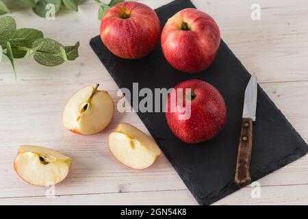 In einer rechteckigen schwarzen Schieferschale auf einem weißen Holztisch befinden sich drei saftig schöne reife rote Äpfel der frühen Wintersorte Gala. Neben dem slic Stockfoto