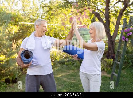 Aktives reifes Paar, das mit Yogamatten steht und sich nach dem Training im Garten im Freien fünf hoch gibt Stockfoto