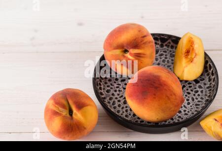 Drei frische, reife, orange-saftige Pfirsiche und ihre Scheiben liegen auf einem schwarzen Tonteller auf einem weißen Holztisch Stockfoto