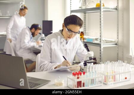Junge Wissenschaftlerin, die sich Notizen gemacht hat, während sie im modernen mikrobiologischen Labor arbeitet Stockfoto