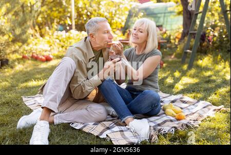 Ein romantisches, älteres Paar, das im Garten picknickt, auf der Decke sitzt und Toast isst und Zeit im Freien verbringt Stockfoto